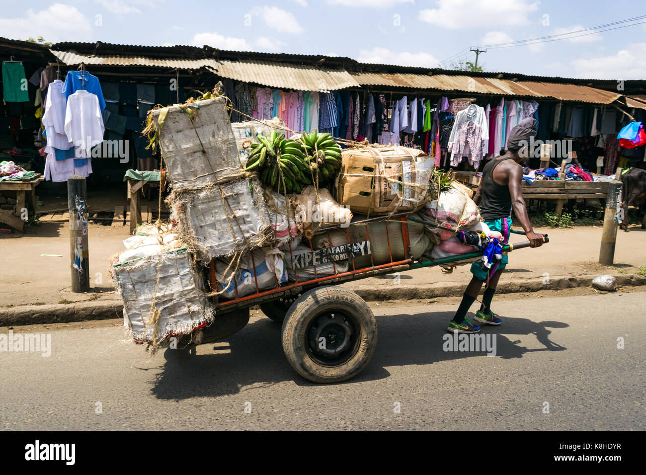 A man pulls a handcart with goods on along road with clothes stalls on pavement in background, Nairobi, Kenya Stock Photo