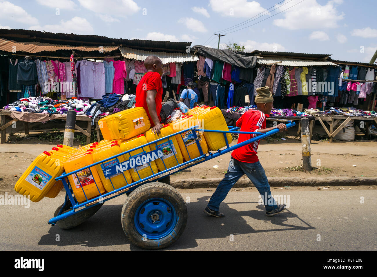 Two men pull a handcart with fresh water containers on road with people and clothes stalls on pavement in background, Nairobi, Kenya Stock Photo