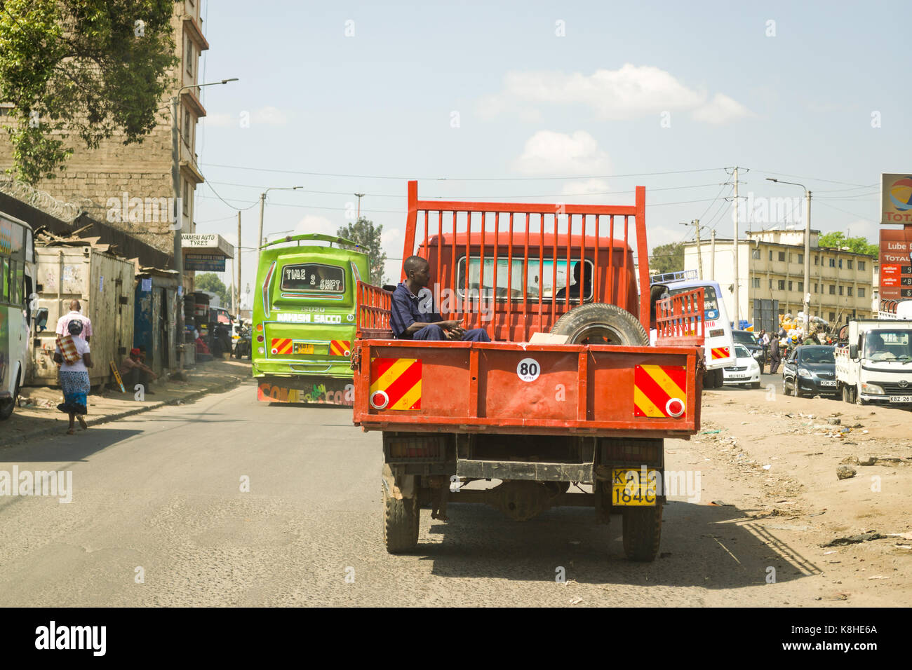 Man sits in back of small open truck as it drives along road with other vehicles on, Nairobi, Kenya Stock Photo