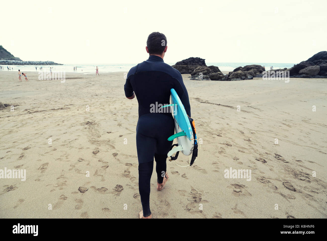 Rear view of man in wetsuit carrying surfboard while walking on beach Stock Photo