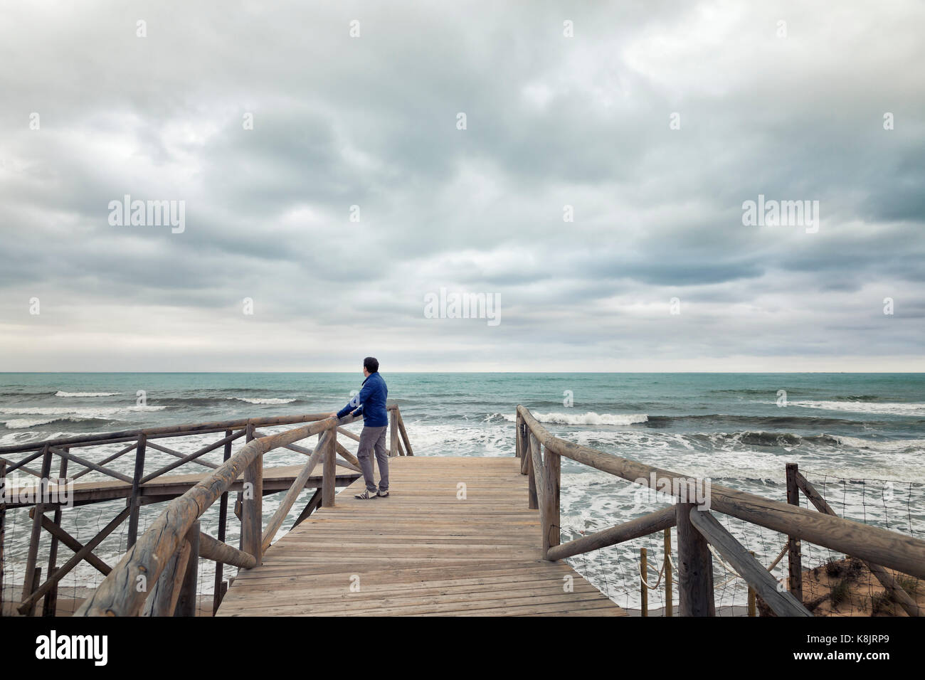 man on boardwalk looking at the sea in a clouded day. Guardamar beach. Alicante, Spain Stock Photo