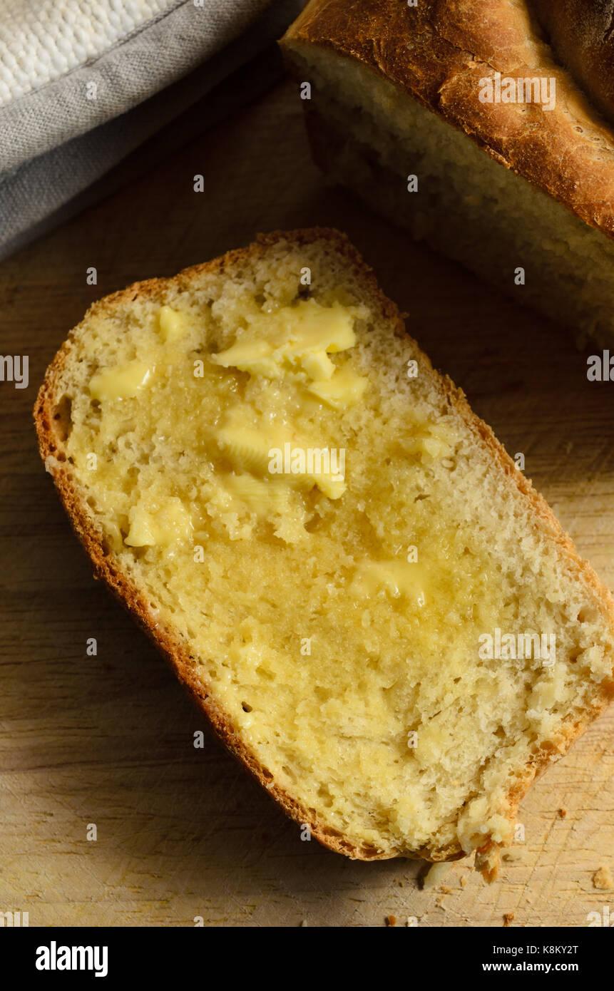 Overhead shot of a slice of freshly baked, warm bread with butter melting into surface. On wooden board with oven gloves, loaf and crumbs. Stock Photo