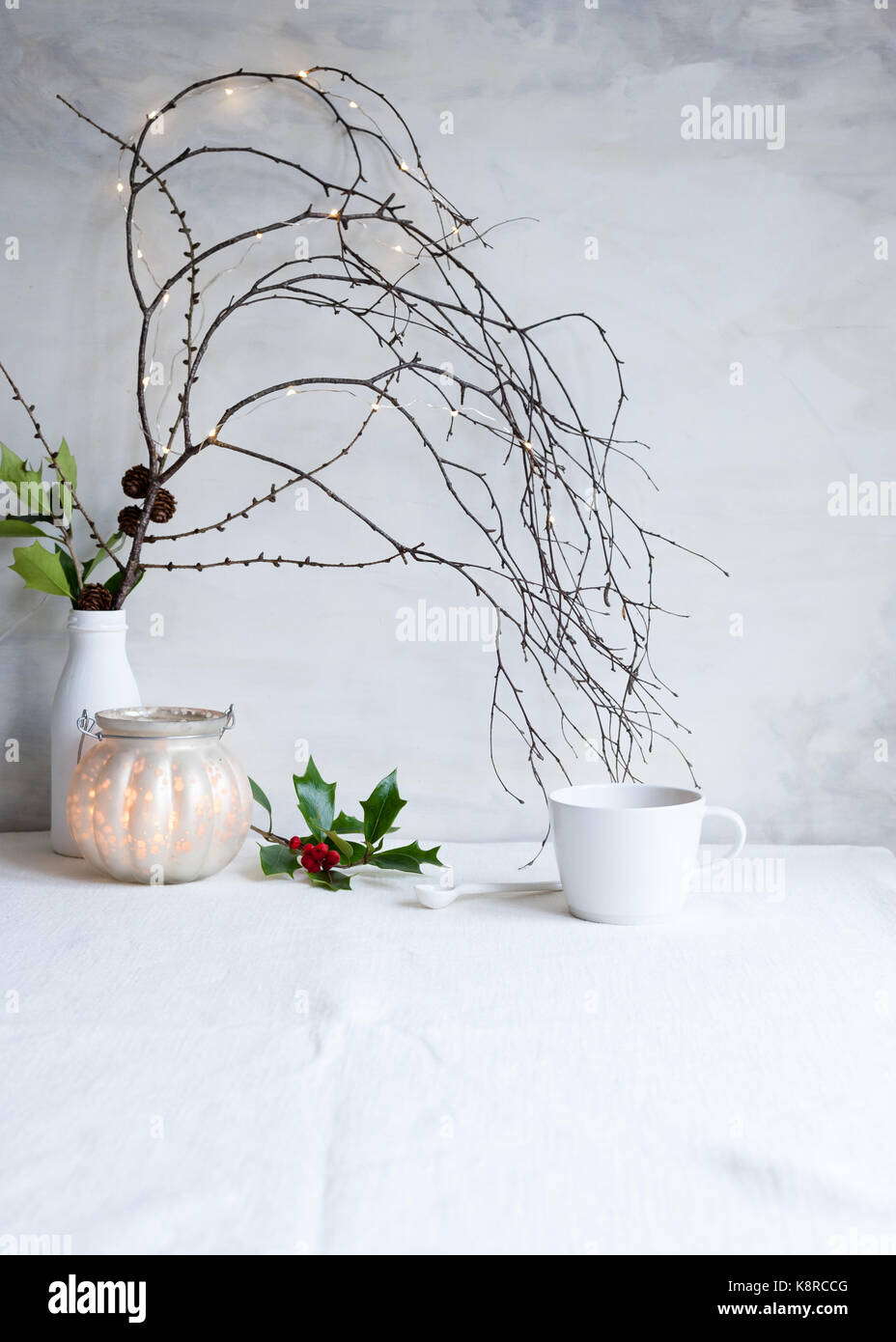 A simple Christmas arrangement with branches, twigs, fairy lights, pine cones and holly, a lit candle, and a teacup. Styled still life, natural light Stock Photo