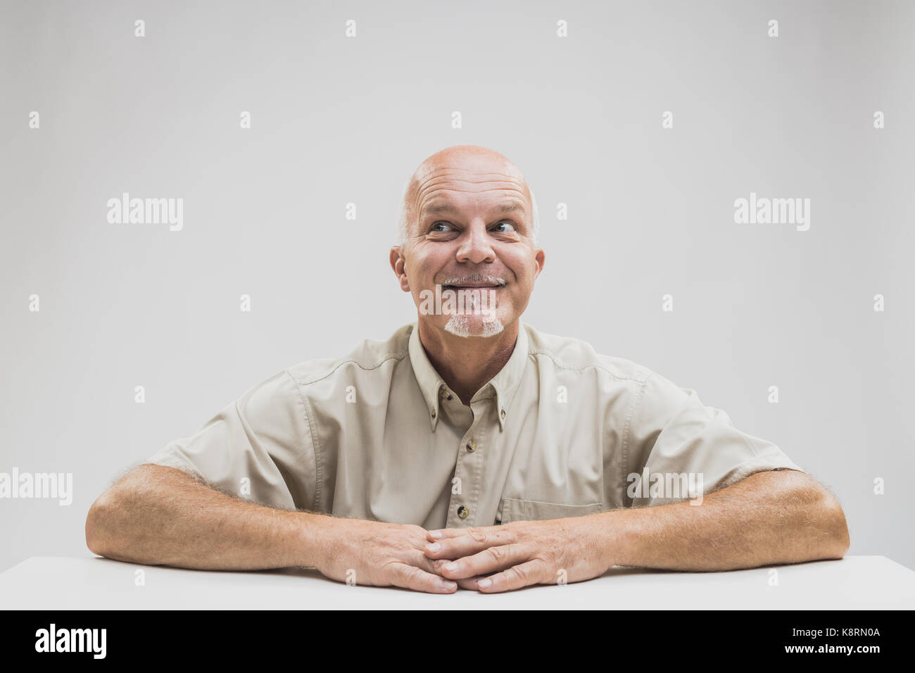 Senior man seated at a table looking up with a smile and a look of gleeful anticipation Stock Photo