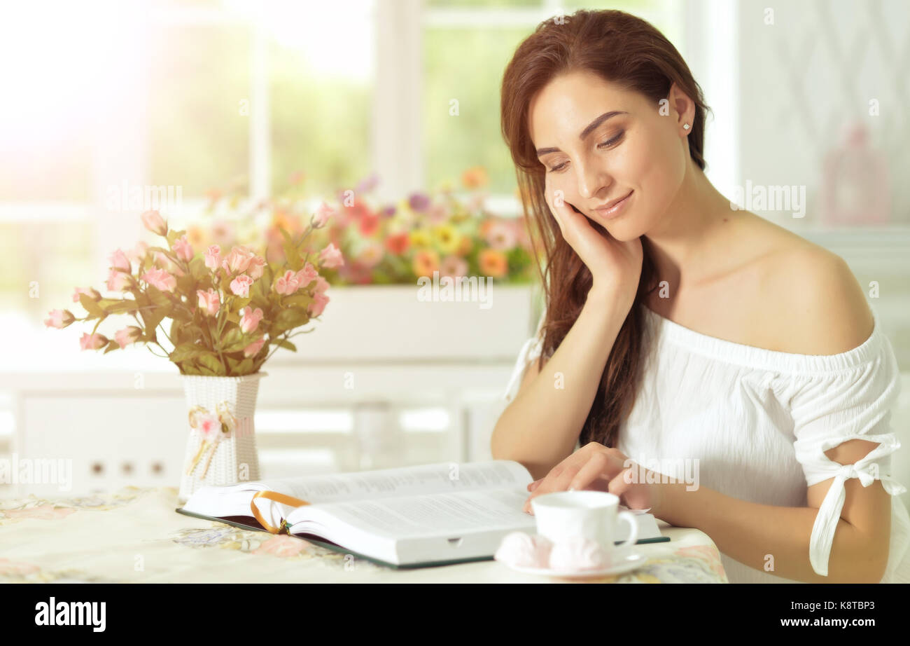 Beautiful young woman sitting at table and reading interesting book ...