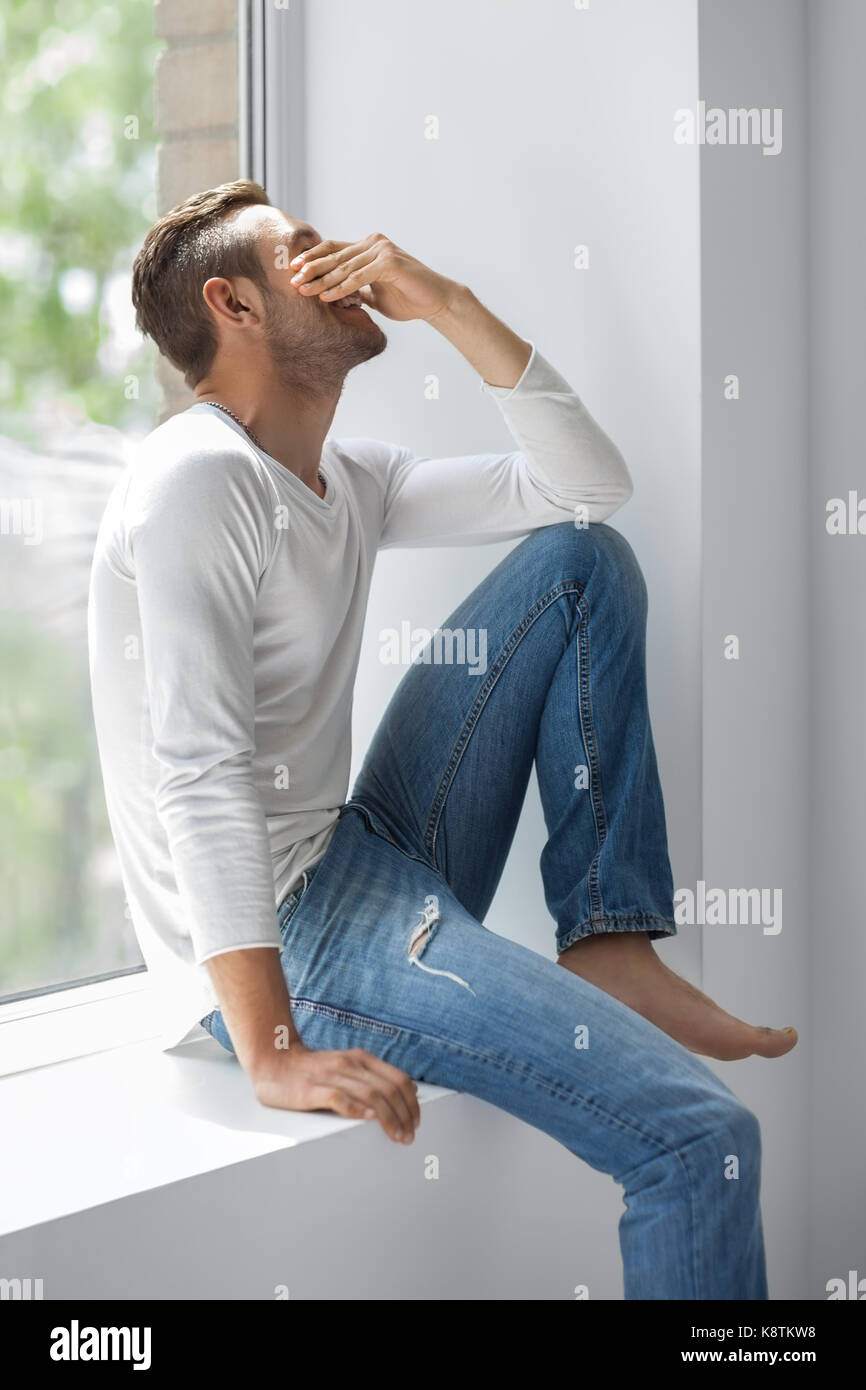 Handsome shy man close face with hand sitting on window sill Stock Photo