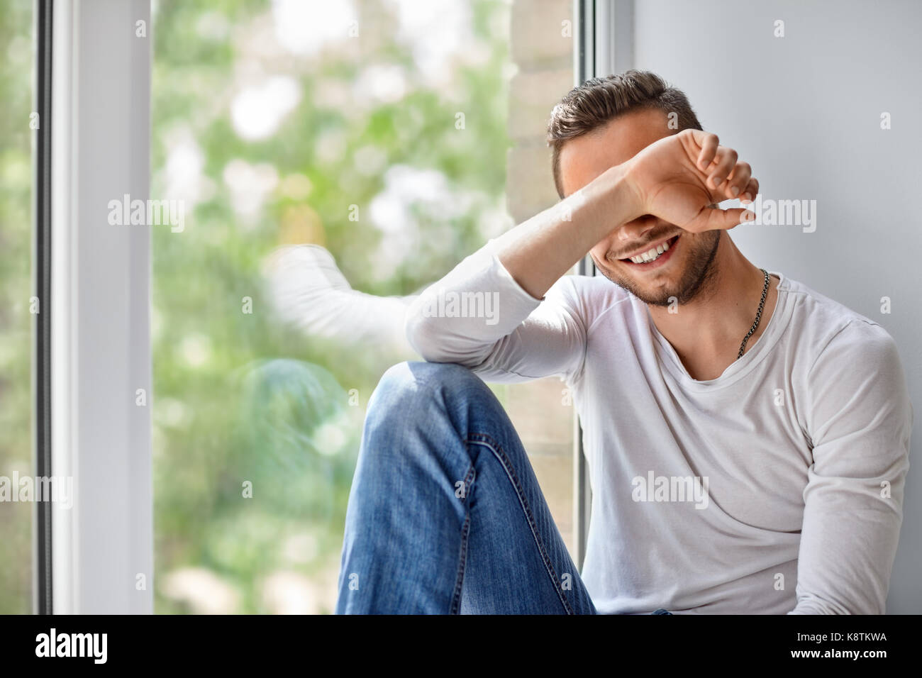 Smiling shy man close face with hand sitting on window sill Stock Photo