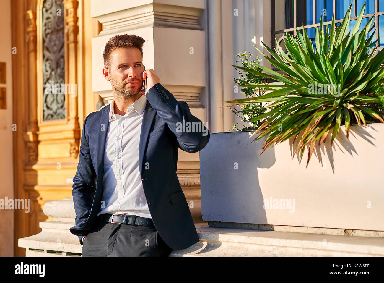 A handsome young businessman leaning against the wall at the entrance of a building while talking on his phone in the sunshine Stock Photo