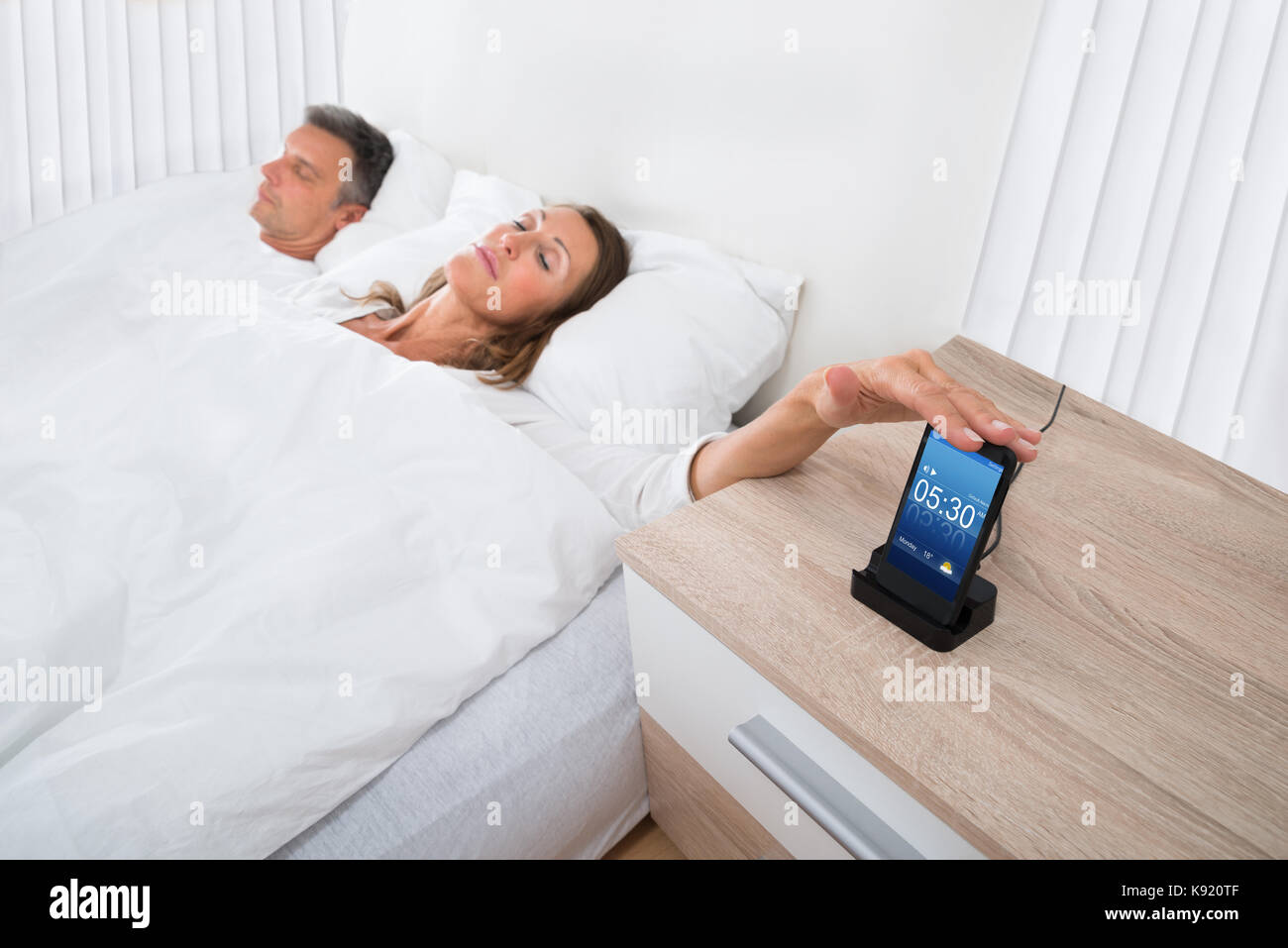 Woman On Bed Snoozing Alarm Clock On Cell Phone Screen While Sleeping Stock Photo