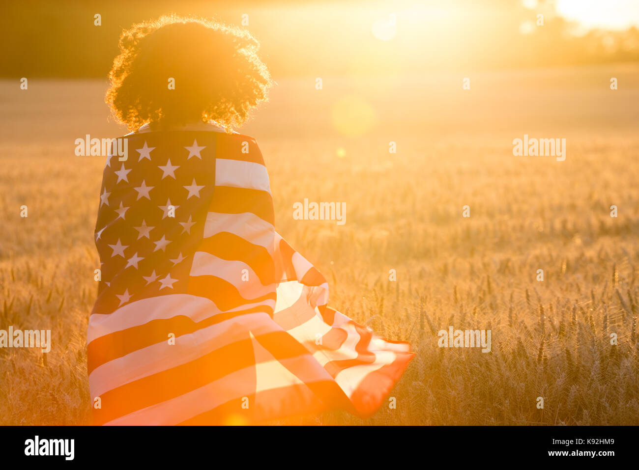 Mixed race African American girl teenager female young woman in a field of wheat or barley crops wrapped in USA stars and stripes flag in golden sunse Stock Photo