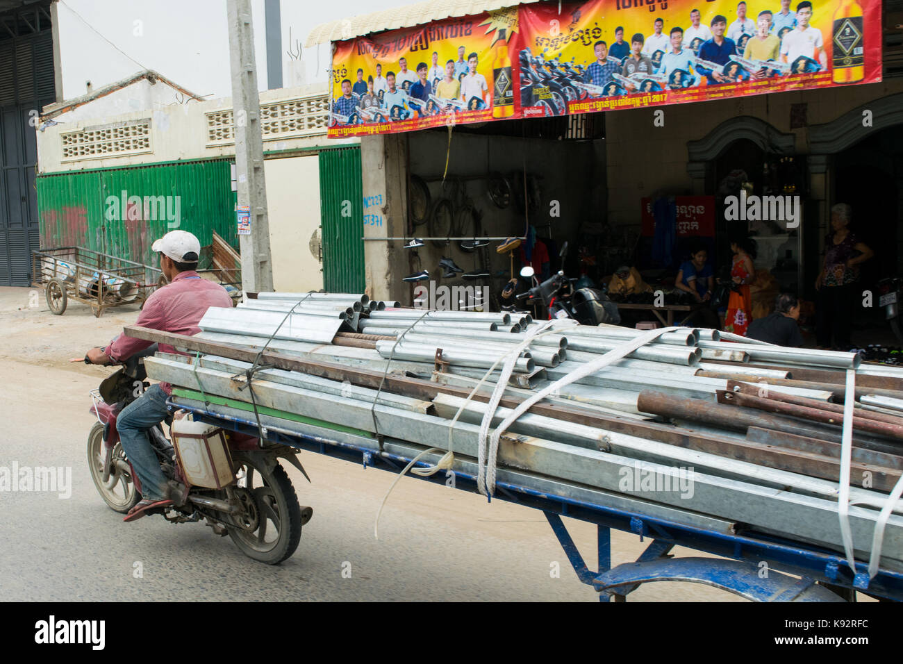 A man driving a motorcycle with an attached trailer loaded with metal pipes. Driving through the busy roads of Phnom Penh, Cambodia, South East Asia Stock Photo