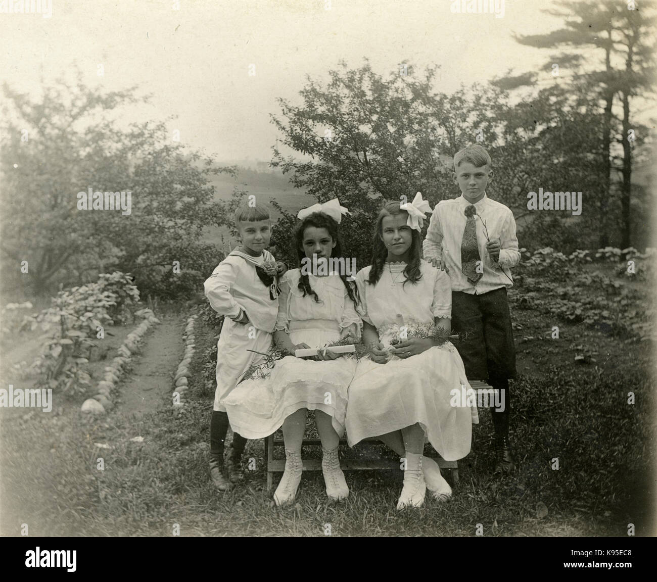 Antique c1920 photograph, two girls in white holding scrolls, possibly on the day of their First Communion; one girl wears a cross pendant. They are flanked by two boys. Location unknown, probably New England. SOURCE: ORIGINAL PHOTOGRAPH. Stock Photo