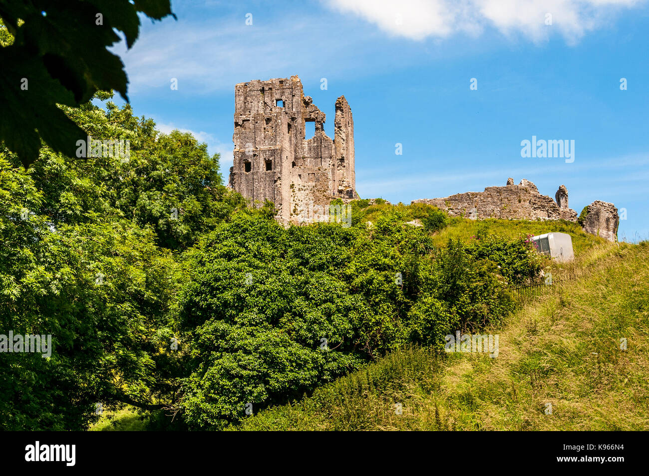 The Keep of the romantic ruins of ancient Corfe Castle standing above a fallen tower and walls on a hilltop above a grassy and wooded steep slope Stock Photo