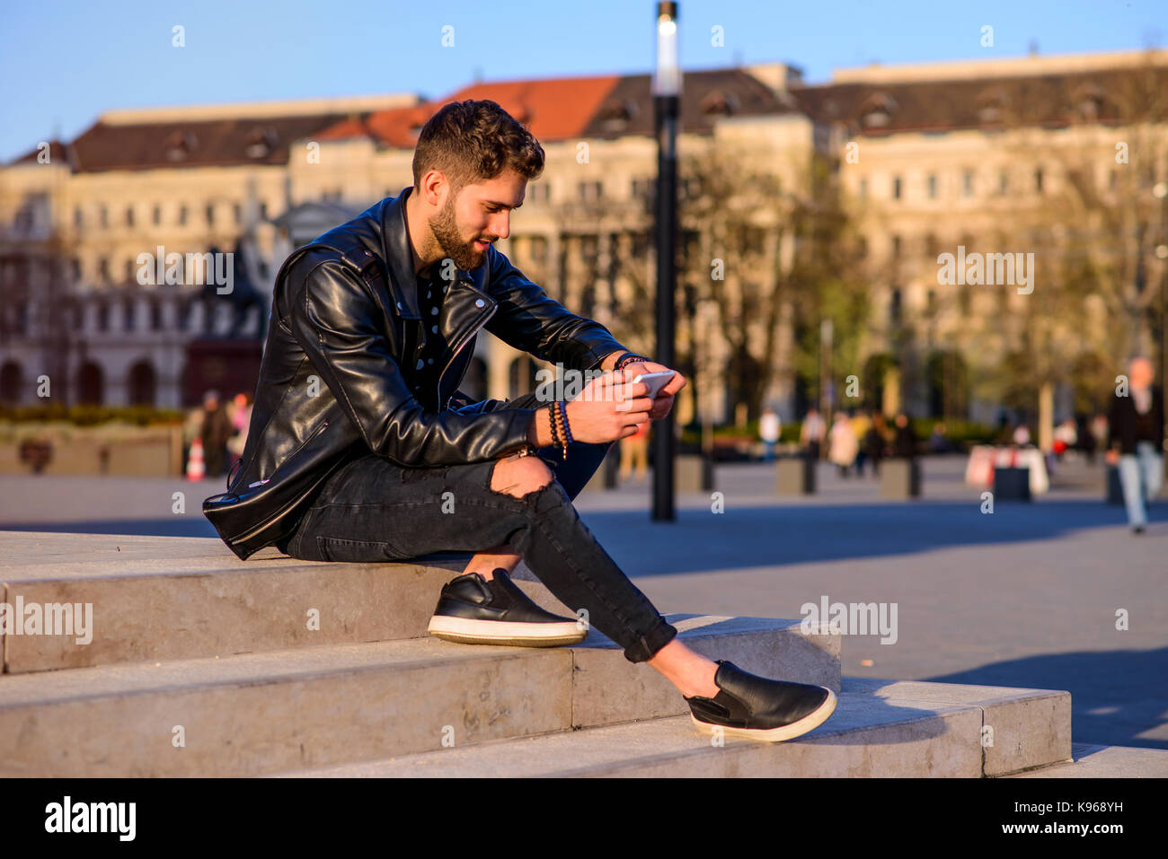 A handsome young hipster man sitting on the stairs on a square while ...