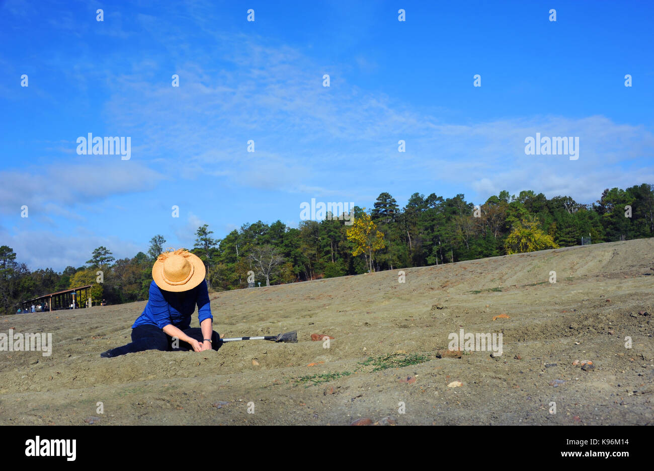 Woman, wearing a straw hat, sits in the dirt field at Crater of Diamonds State Park in Murfreesboro, Arkansas.  She is digging for diamonds. Stock Photo