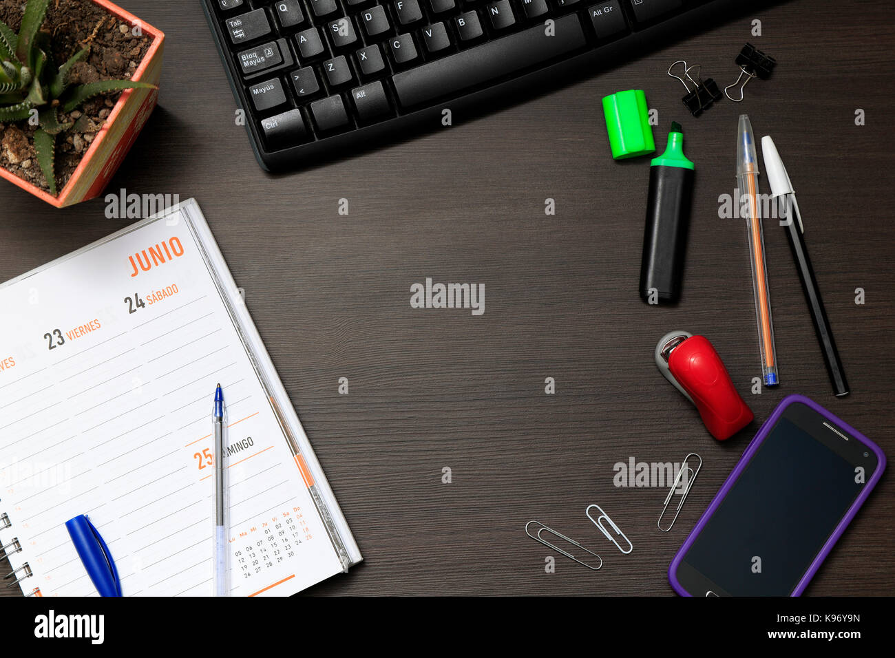 Top view of Office desk with keyboard, diary, clips, pens, highlighter, buckle, a cactus plant, and a cell phone. Stock Photo