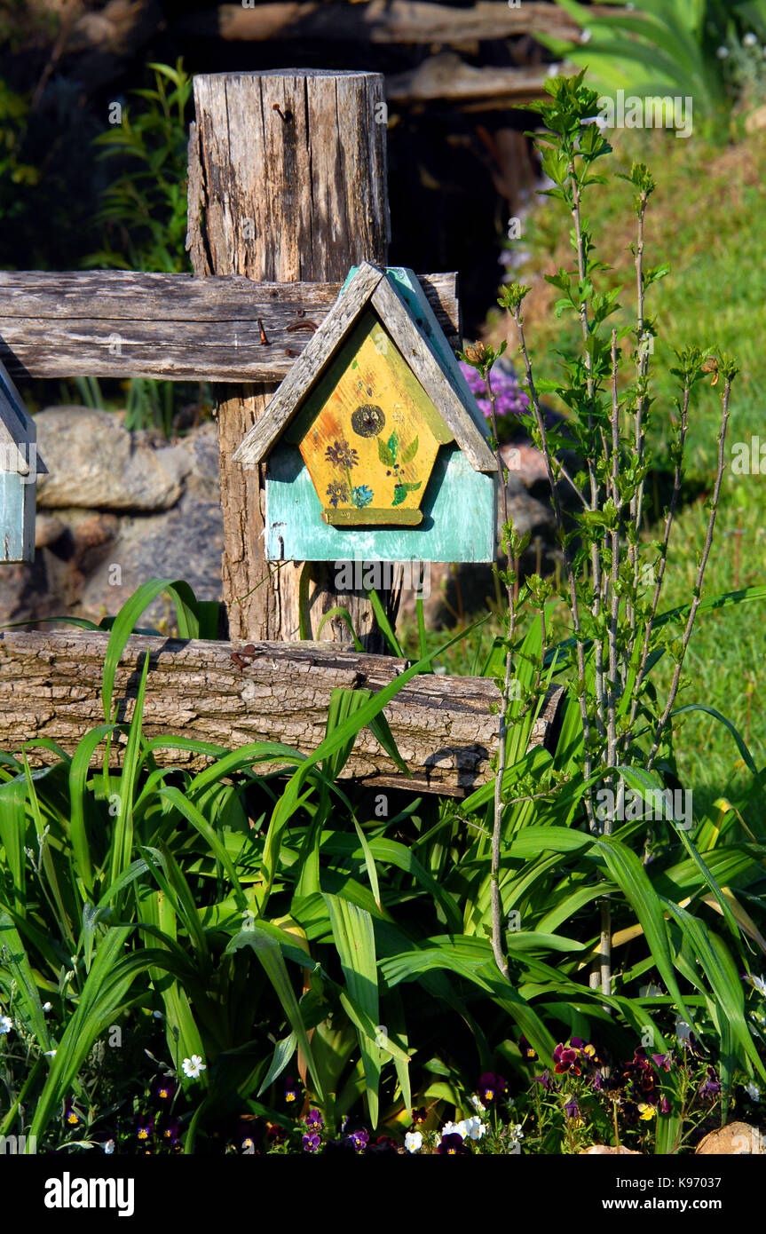Rustic fence is graced with a brightly painted birdhouse.  Flowers bloom in garden setting. Stock Photo