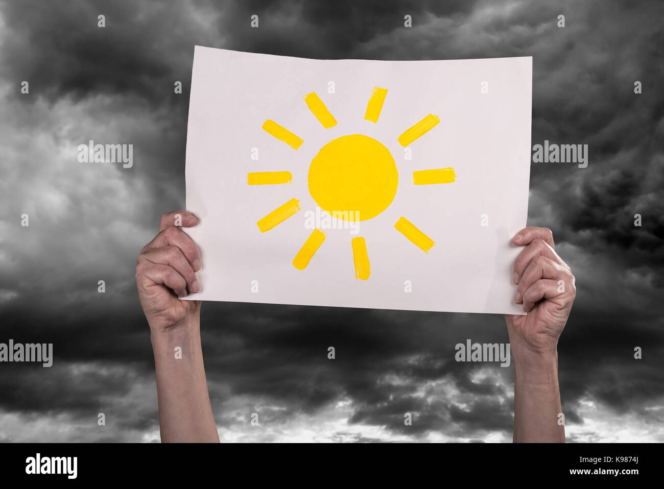 Hands holding a piece of paper with a painted sun high in front of dark clouds. Stock Photo