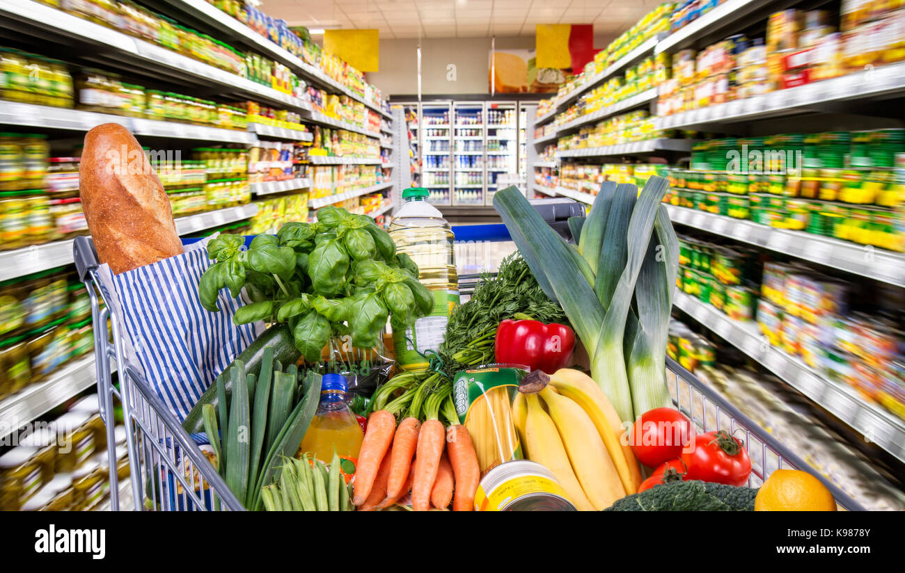 Detail of a shopping cart with fruit and vegetables between goods shelves in the supermarket Stock Photo