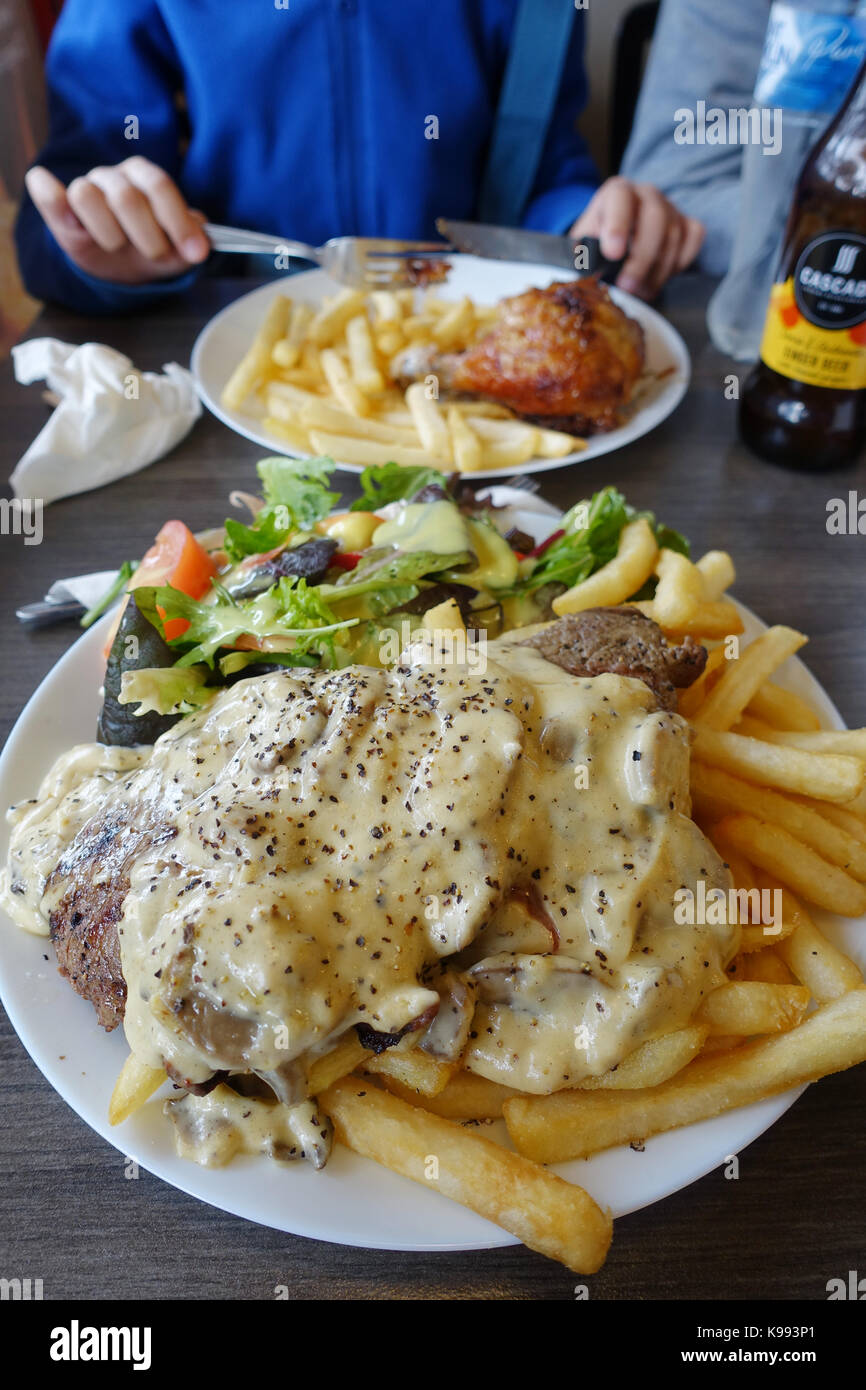 A plate of steak with mushroom sauce and chips with a bowl of Greek salad and Chicken Schnitzel burger in the background Stock Photo