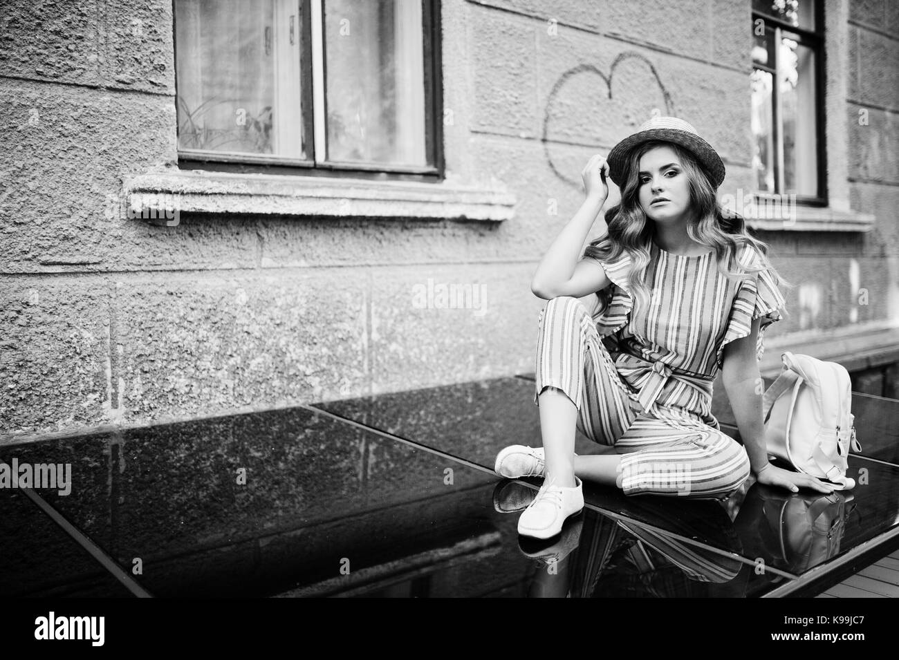 Portrait of a fabulous young woman wearing striped overall and hat sitting on black shiny surface with backpack next to the building. Black and white  Stock Photo