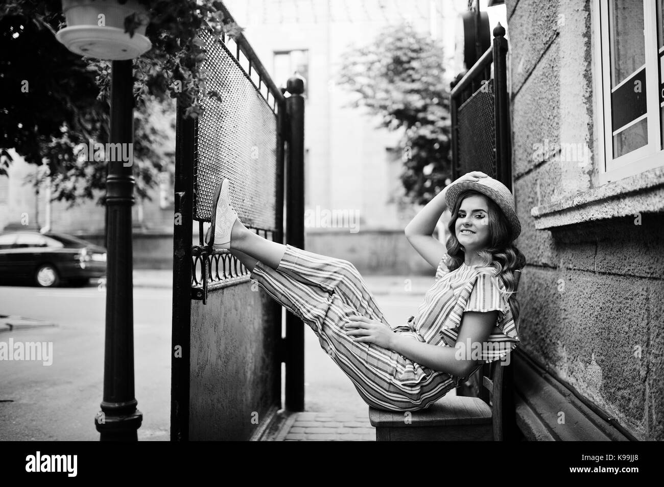 Portrait of a fabulous young woman in striped overall and hat sitting and posing on the chair outdoor. Black and white photo. Stock Photo