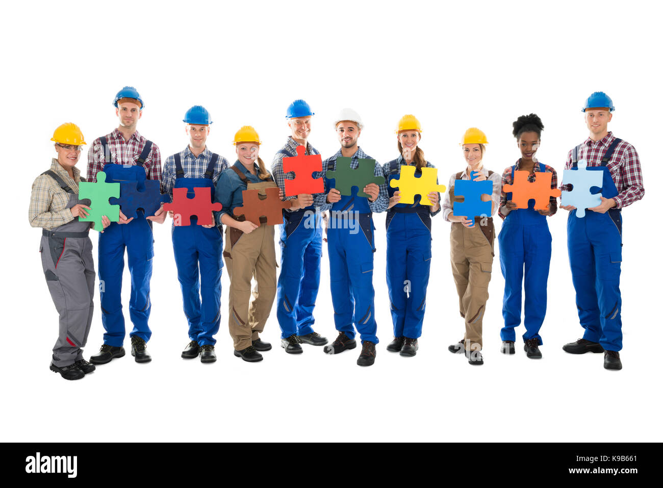 Portrait of confident construction workers holding jigsaw pieces while standing in row against white background Stock Photo