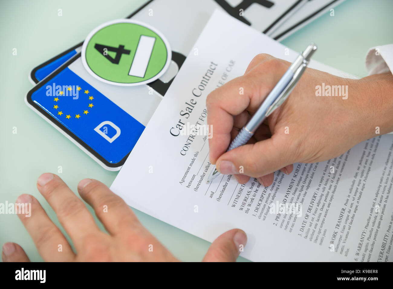 Close-up Of A Person Filling Car Sale Contract Form With Number Plate On Desk. Contract Paper Contains Placeholder Text Stock Photo