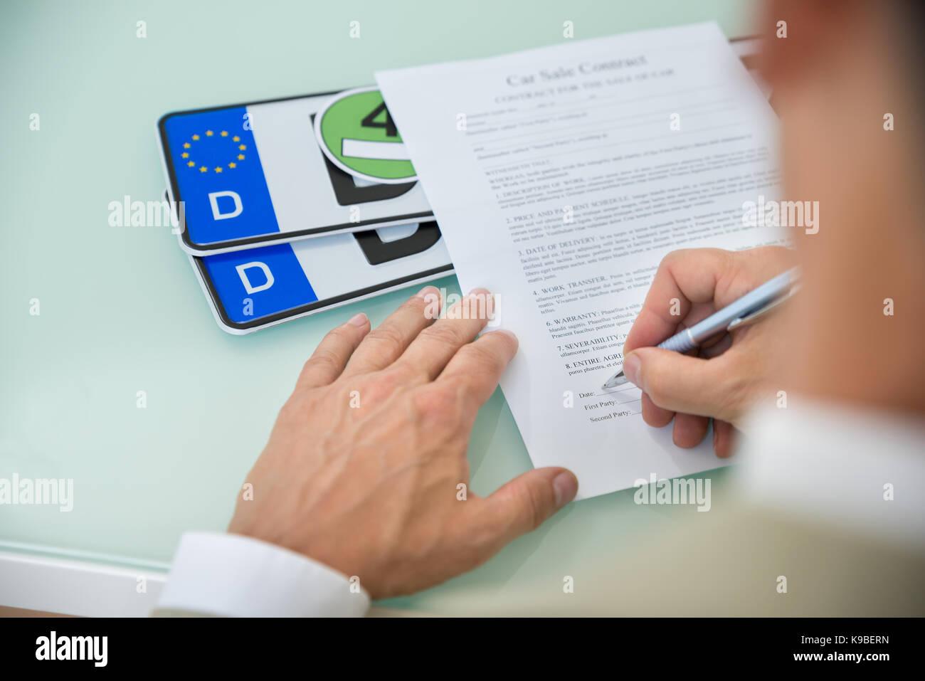 Close-up Of A Businessman Filling Car Sale Contract Form With Vehicle Registration Plate On Desk. Contract Paper Contains Placeholder Text Stock Photo