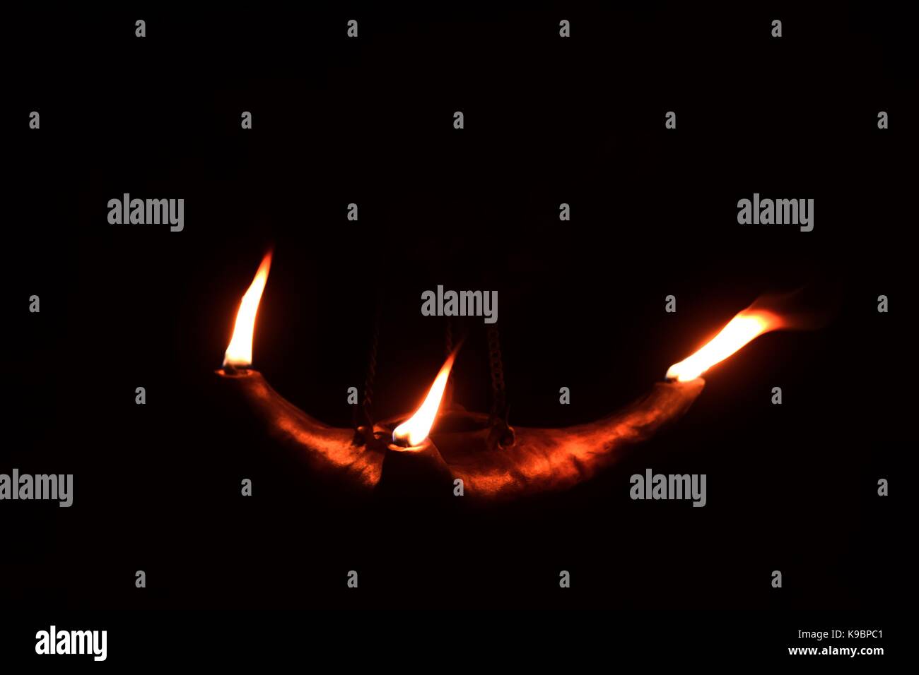 Three flames coming out of a clay oil lamp, in the dark. The lamp is suspended by a metal chain. Shot in Malta Stock Photo