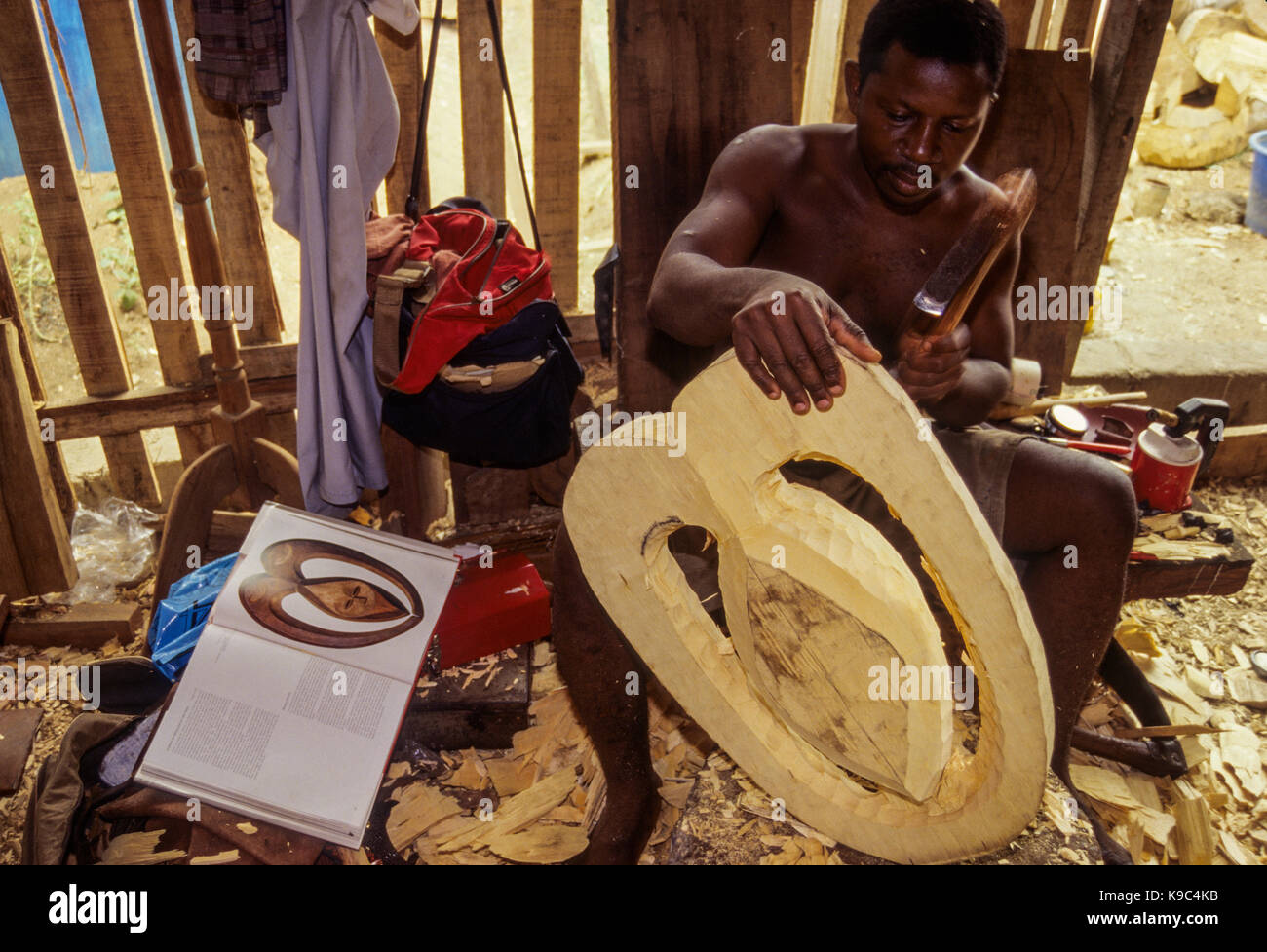 Abidjan, Ivory Coast, Cote d'Ivoire.  Using an African Art Book for Reference this Ghanaian Carver Makes a Replica of a Kwele Mask, a People Living on the Borders of Cameroon, Gabon, and Congo. Stock Photo