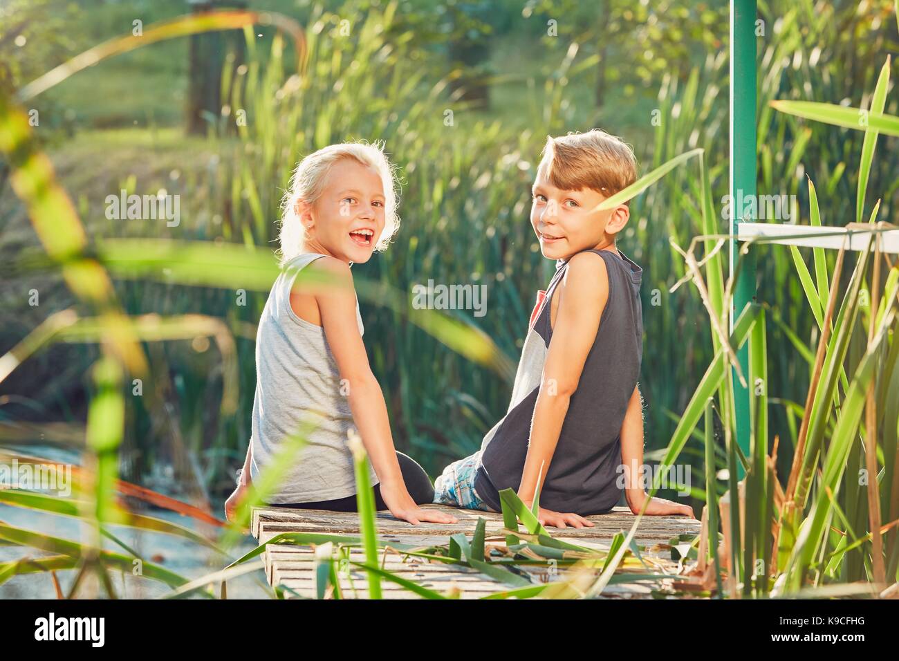 Summertime in the countryside. Two children (sibling or best friends) sitting on the pier of the lake. Stock Photo
