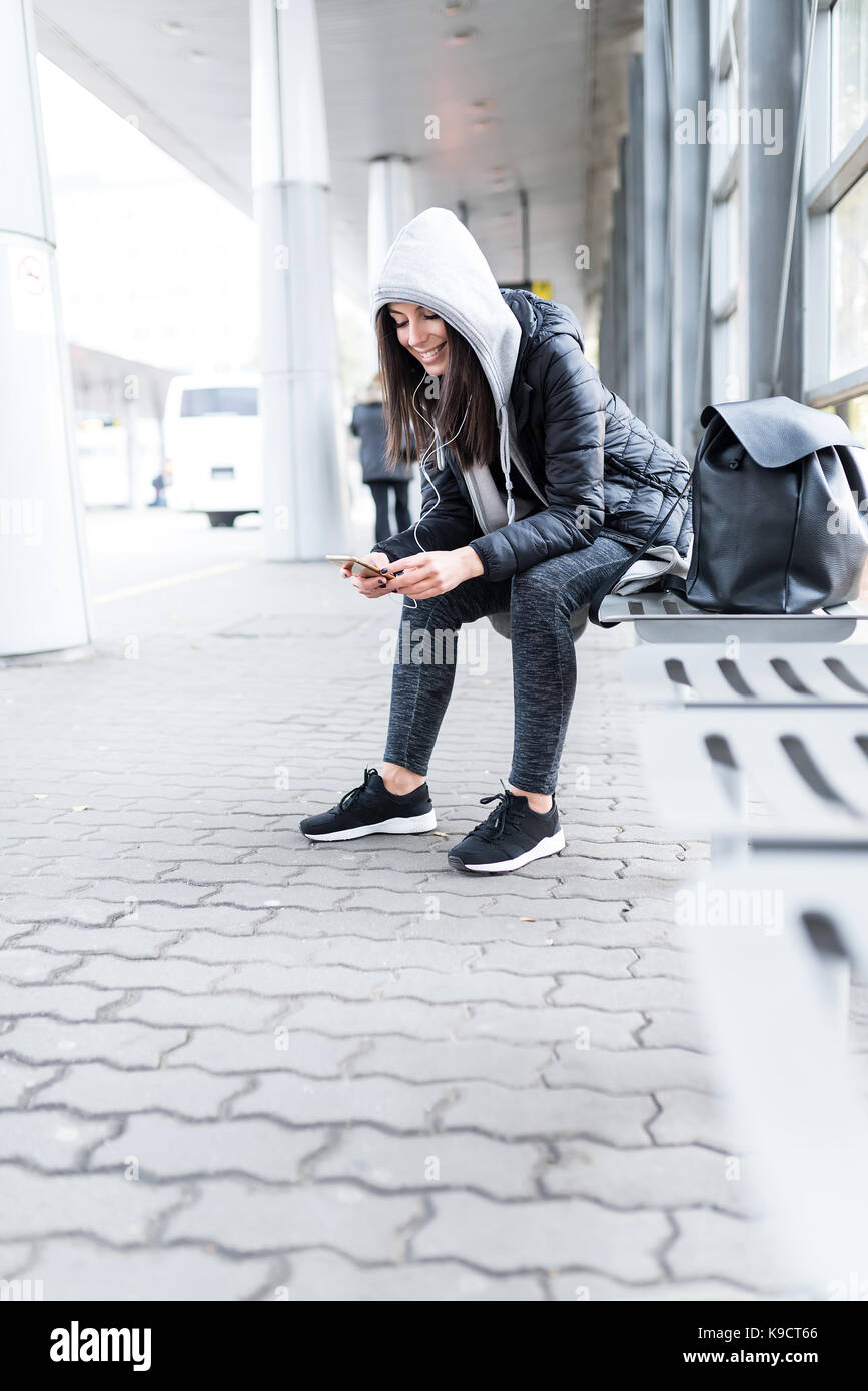 Beautiful young woman waiting for the Bus at a Bus stop while listening to  music on her smartphone Stock Photo - Alamy