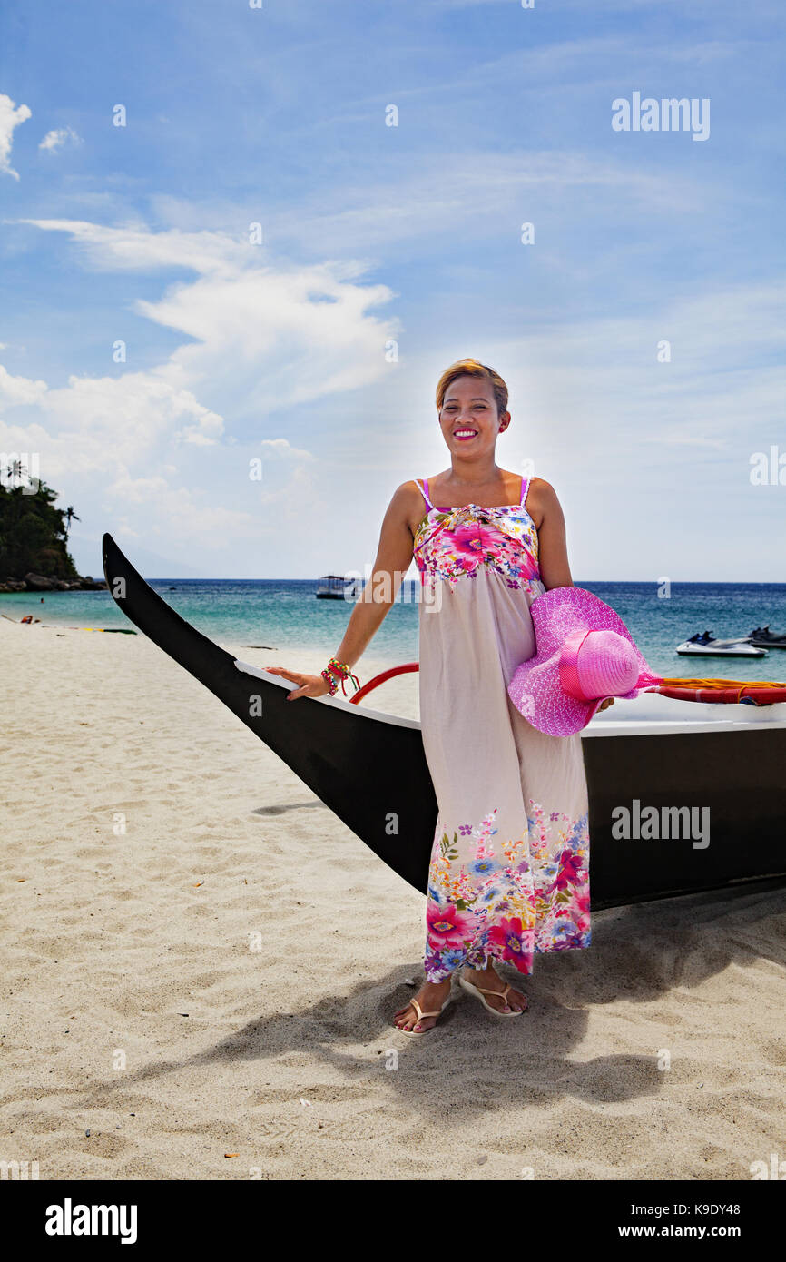 A beautiful Filipino woman poses by an outrigger in her flowery dress and pink wide-brim hat on White Beach near Puerto Galera, Mindoro, Philippines. Stock Photo