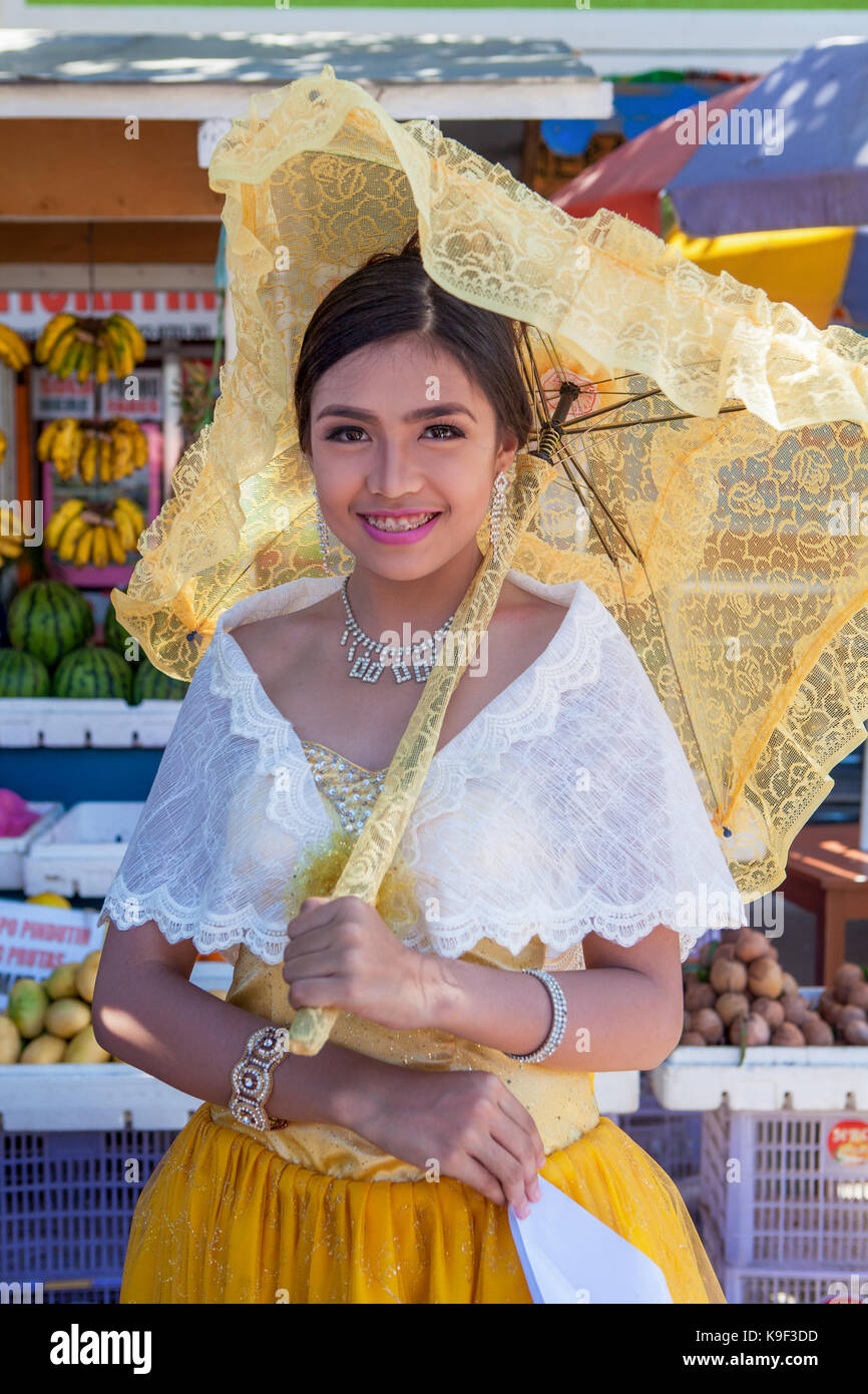 A beautiful Filipino high school teenage schoolgirl is dressed in the Spanish style wearing a yellow dress, white lacey shawl and a lacy, yellow paras Stock Photo