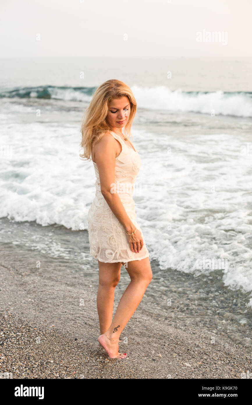 Full length vertical portrait of a blonde woman barefoot and wearing short  beige cocktail dress by the sea Stock Photo - Alamy