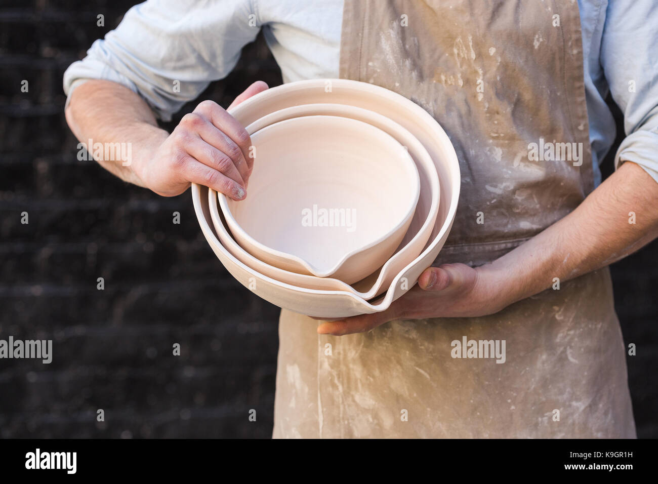 potter, utensils, ceramics art concept - close-uo on male hands holding some beautiful bowls, cute finished clay products and craftsman in the apron on background of dark gray brick wall. Stock Photo