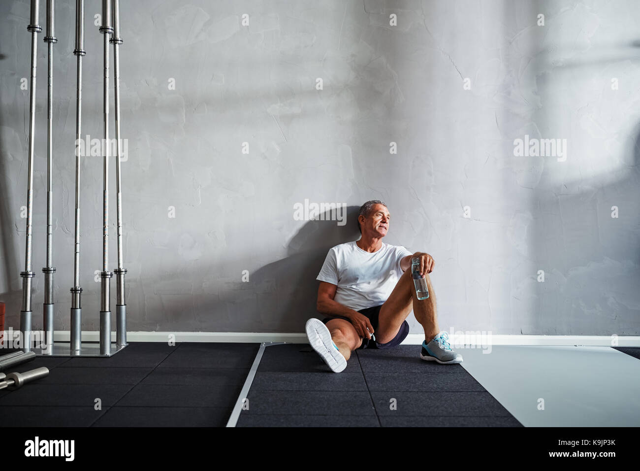 Senior man sitting on the floor of a gym drinking from a water bottle ...