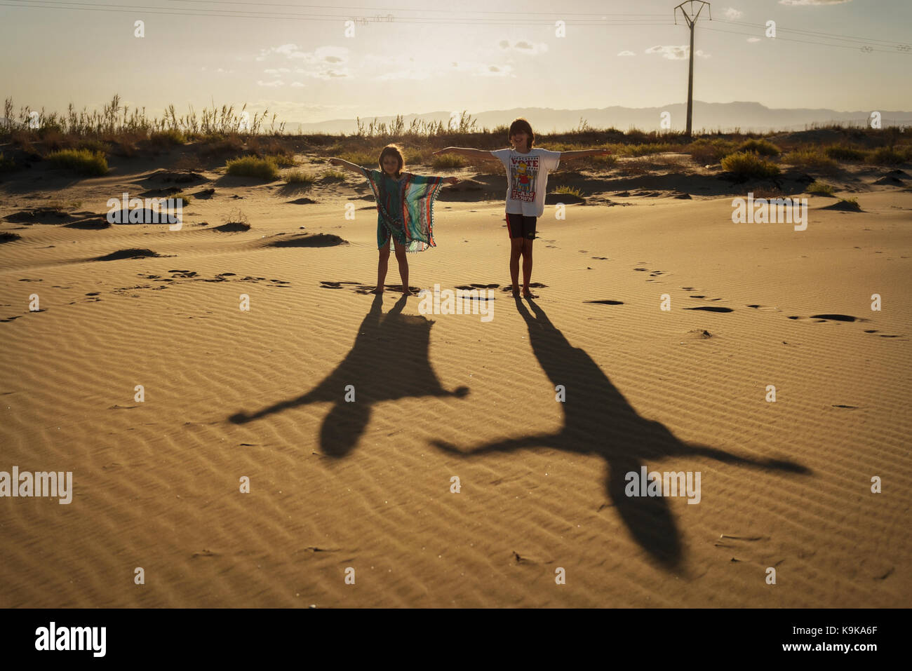 Boy, girl and their shadows standing in on a sandy beach with outstretched arms Stock Photo