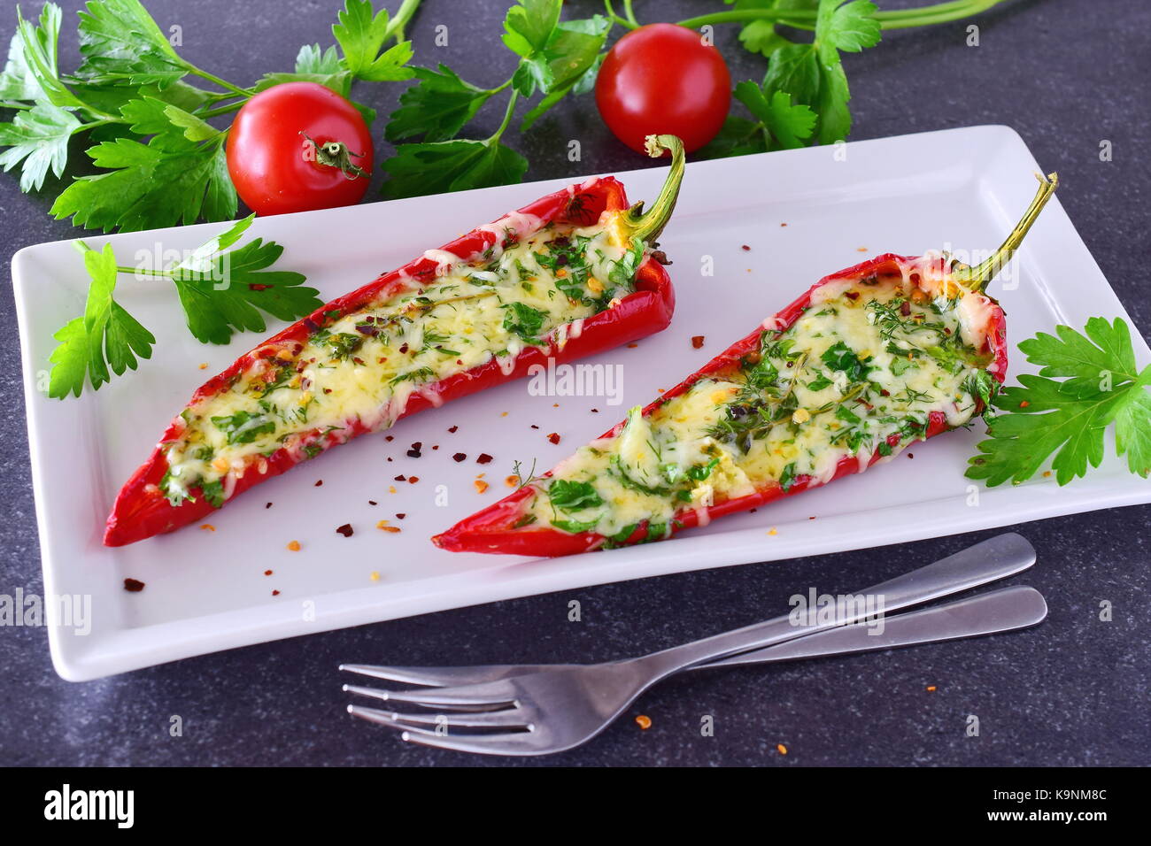 Oven cooked red paprika stuffed with cheese, garlic and herbs on a white plate with parsley and cherry tomatoes an abstract grey background. healthy e Stock Photo