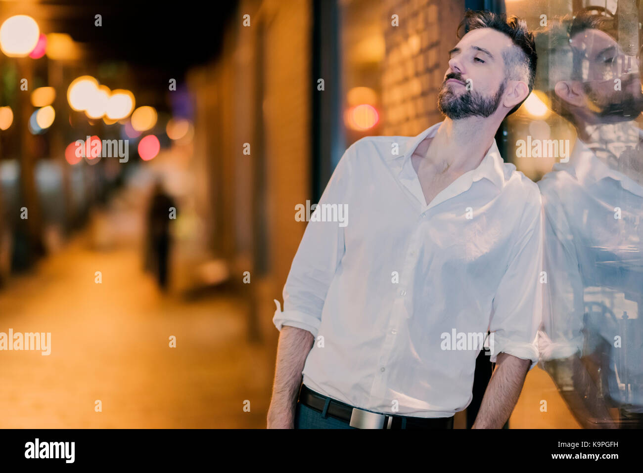 Handsome caucasian male professional photographed on the street at night leaning against a shop window looking down. Stock Photo