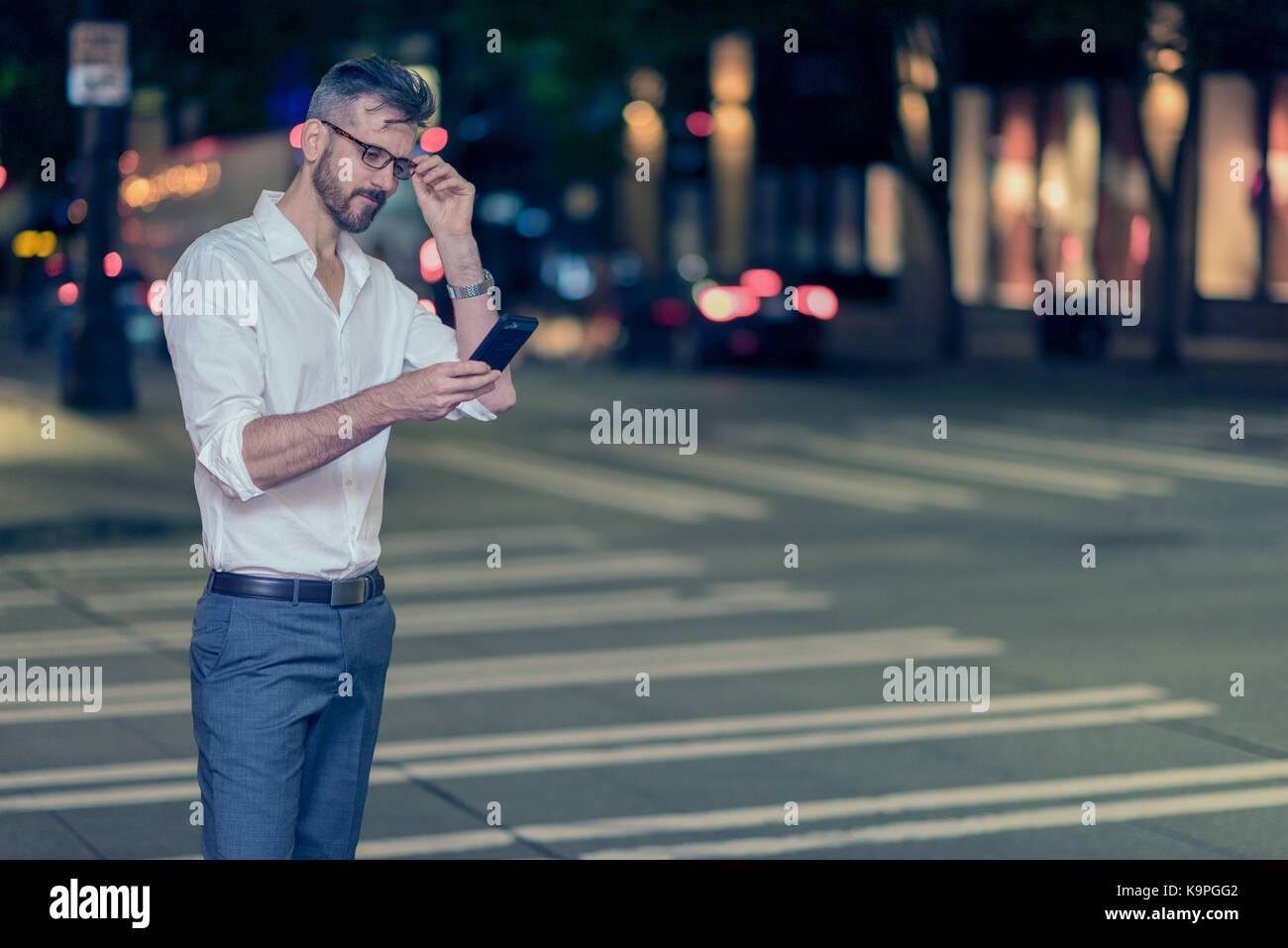 Handsome caucasian male professional photographed on the street at night looking at mobile phone standing at intersection Stock Photo