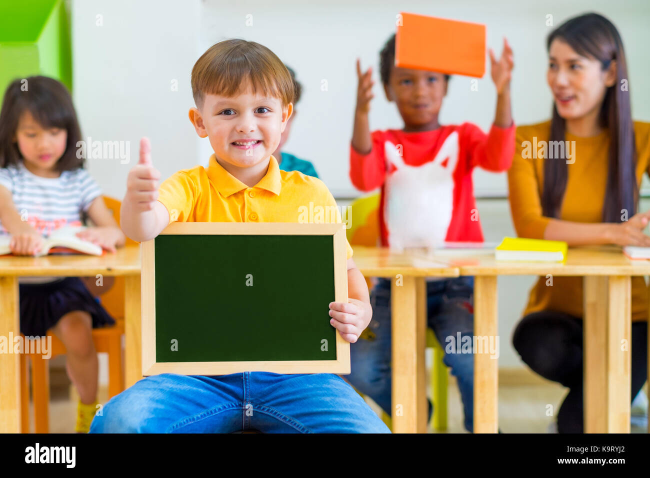 Kid boy thumbs up and holding blackboard with back to school word with diversity friends and teacher at background,Kindergarten school,mock up chalkbo Stock Photo