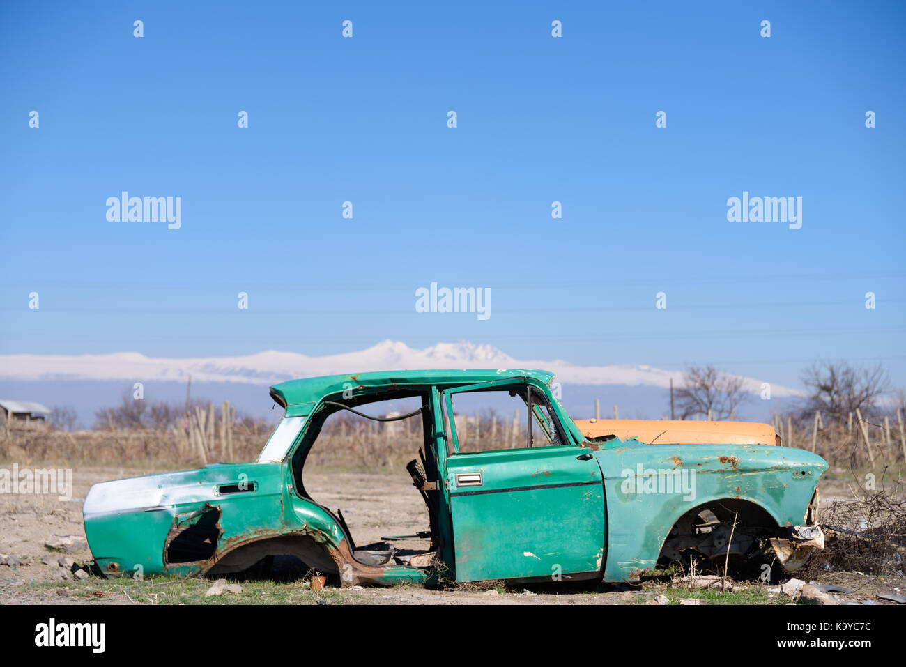 Abandoned and rusty wreck of an old green vintage Soviet Russian car in the middle of dry agricultural land with scenic snow-topped mountains and clea Stock Photo