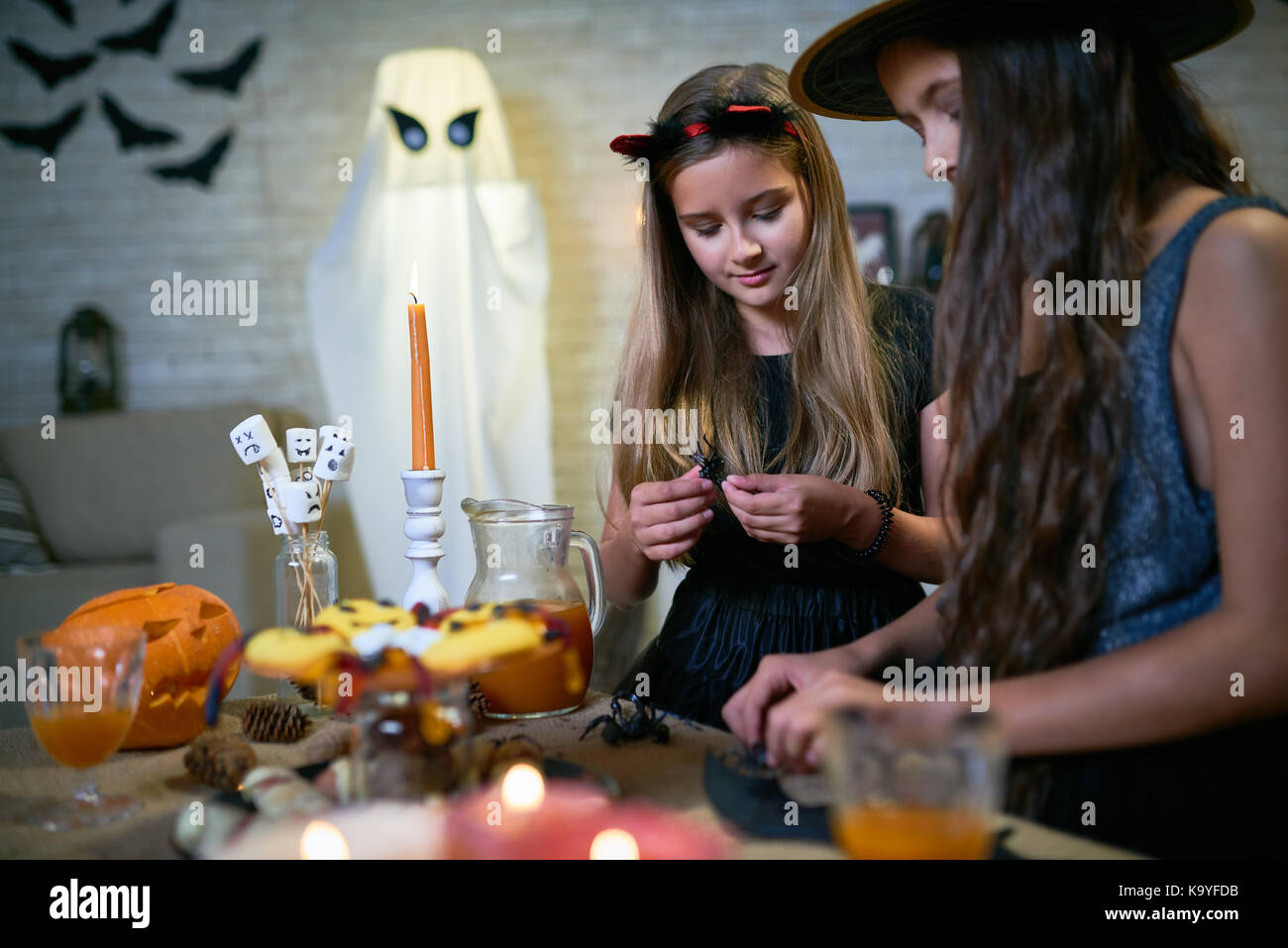 Serious curious girls playing with Halloween toys at table with candies and talking about Halloween traditions or making crafts at kids party Stock Photo