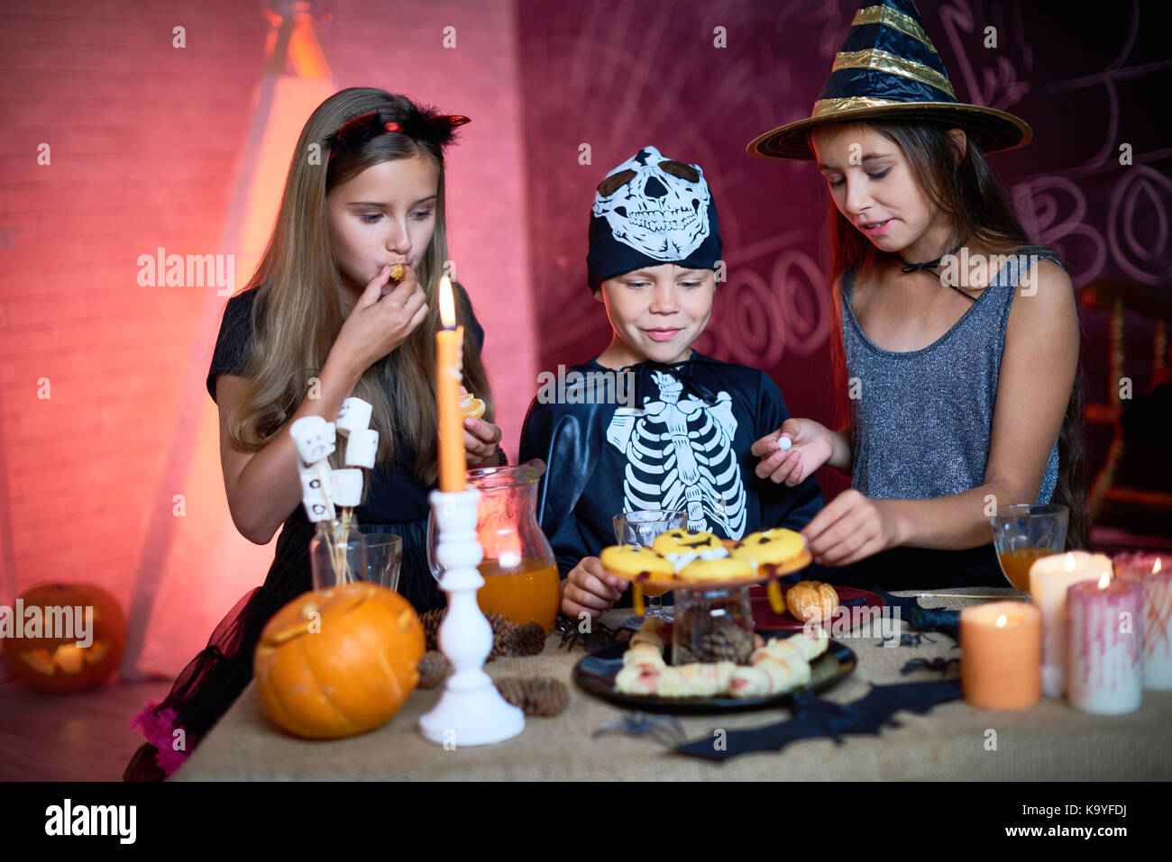 Positive curious kids eating sweet food from candy bar tasting Halloween treats in decorated studio at party Stock Photo