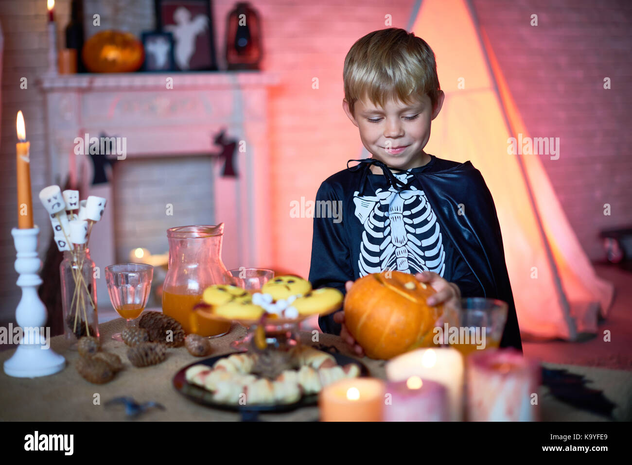 Happy cute blonde boy in skeleton costume with cape taking pumpkin from candy bar table while standing alone in decorated studio for parties Stock Photo