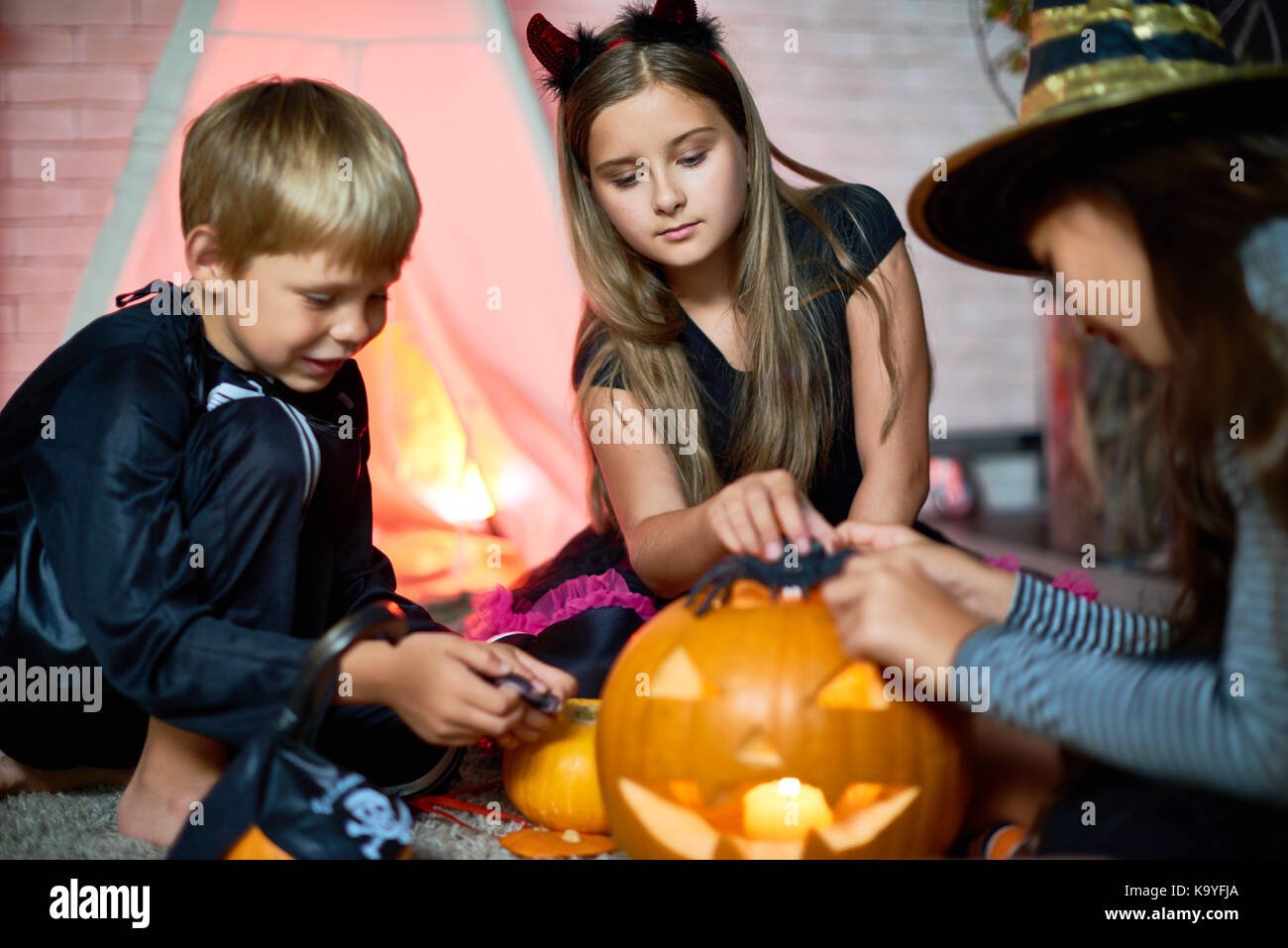 Curious concentrated friends sitting on floor and enjoying game with Halloween toys such as jack-o-lantern and spiders telling scary stories to each o Stock Photo