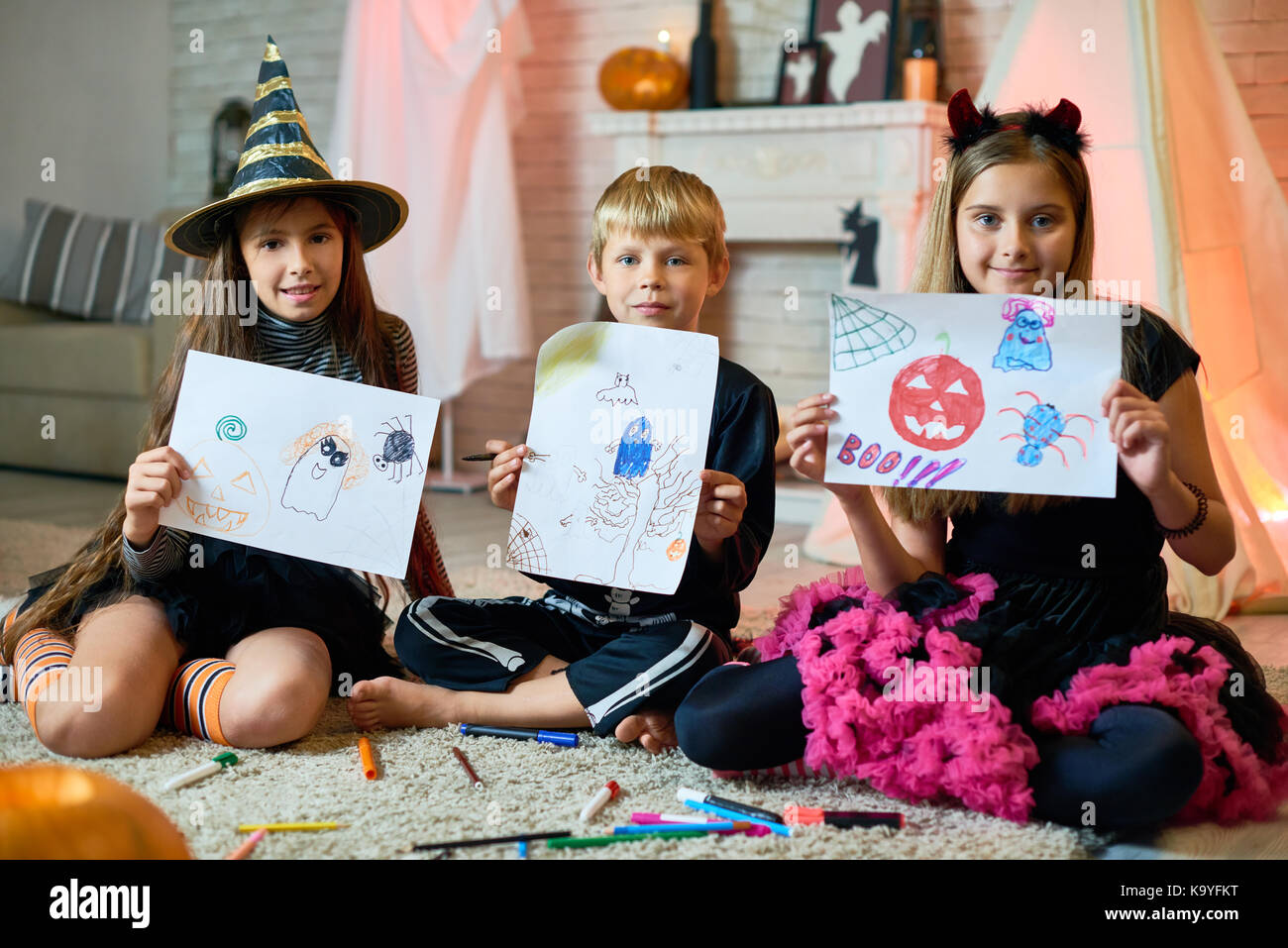 Positive cute friends in Halloween costumes sitting on carpet with splattering markers and showing creative pictures with Halloween characters to came Stock Photo