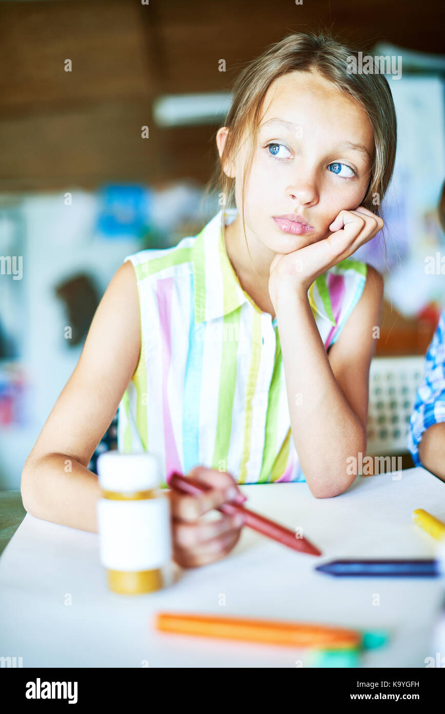Portrait of beautiful little girl daydreaming looking away pensively during art class in school, sitting with blank sheet of paper Stock Photo